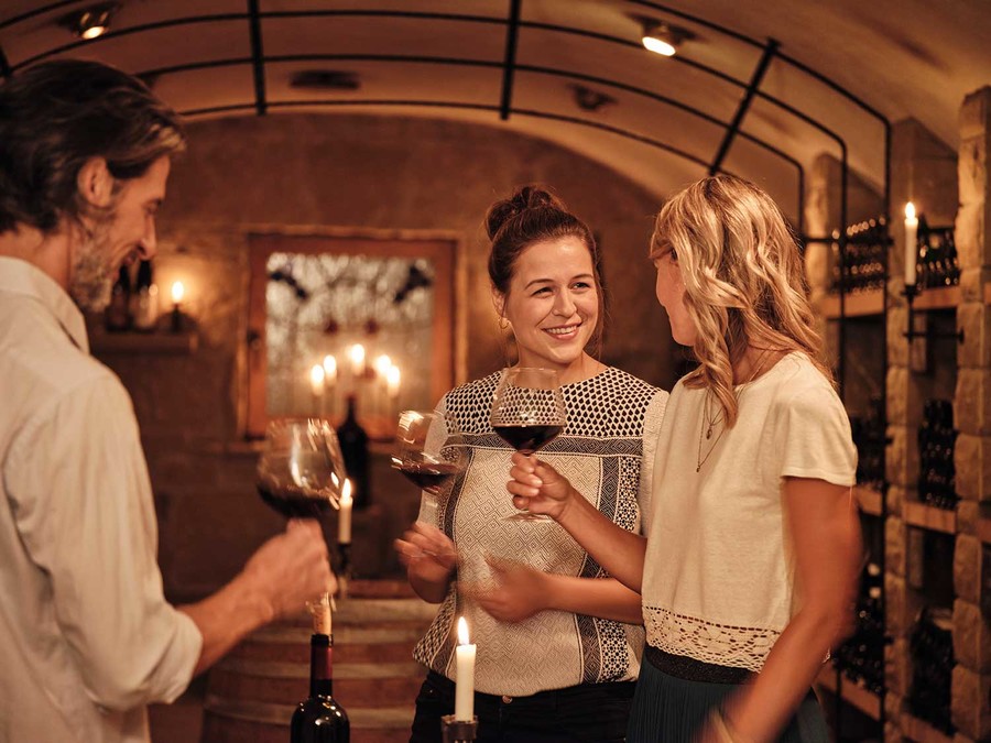 In einem Weinkeller stehen zwei Frauen und ein Mann. Sie halten alle drei ein Weinglas mit Rotwein gefüllt in den Händen. Im Hintergrund stehen Regale mit Wein.