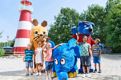 Vor einem rot-weißen Leuchtturm stehen fünf Kinder. Zwischen den Kindern stehen Maskottchen des Ravensburger Spielelands wie zum Beispiel eine orangene Maus, ein blauer Elefant und ein blauer Bär.