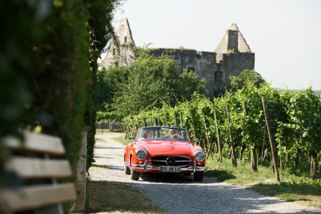 Ein rotes Cabrio fährt auf einem Schotterweg durch Weinberge. Im Hintergrund steht eine alte Burgruine.