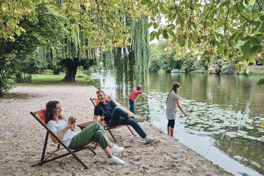 Am Sandstrandufer des Neckars sitzen zwei Personen in Liegestühlen. Zwei Kinder spielen am Ufer im Fluss.