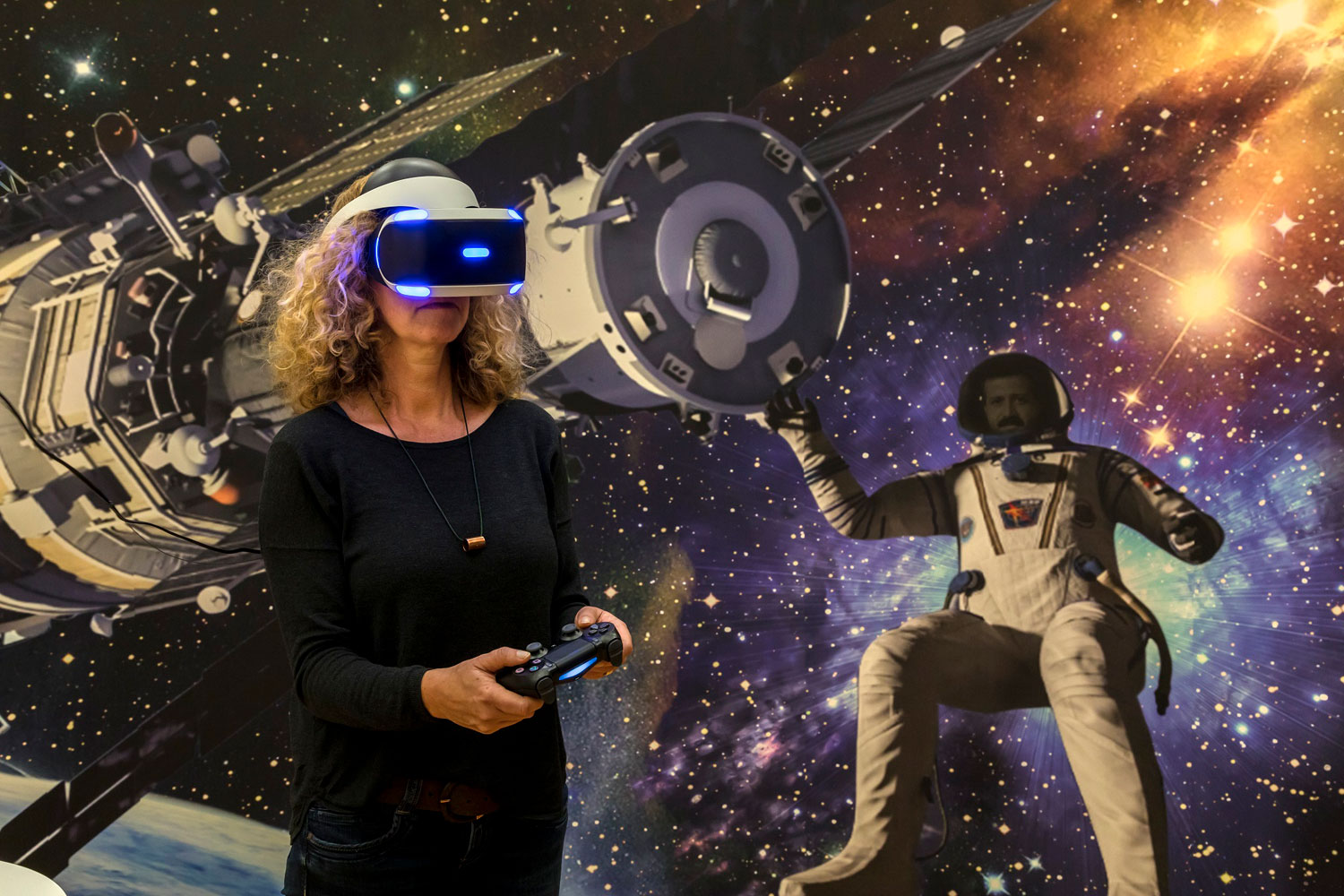 Im Hintergrund ist ein großes Bild aus dem Weltall zu sehen mit Sternen und Kometen darauf.. Links ist eine Raumkapsel und rechts daneben ein schwebender Astronaut in einem Raumanzug. Vor der Wand steht eine Frau, welche eine VR-Brille auf hat. In den Händen hält sie ein länglicher Spiele-Controller.