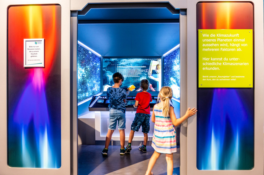 Drei Kinder stehen in einem Eingang und entdecken den Raumgleiter der Klima Arena. Links und rechts sind Infoschilder auf blauem und orangenem Hintergrund angebracht.