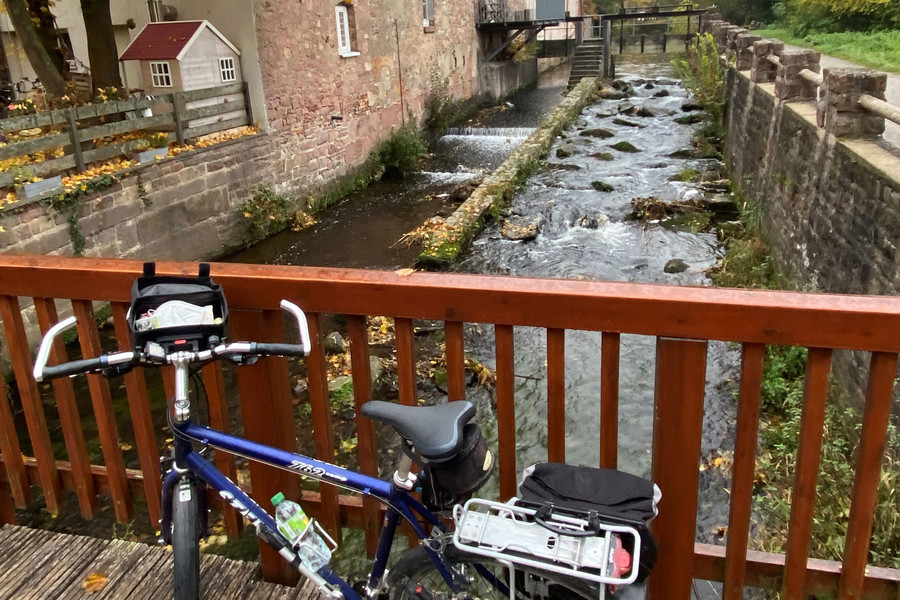 Hinter einem Brückengeländer steht ein Fahrrad mit gepackten Satteltaschen. Unter der Brücke verläuft ein kleiner Fluss.