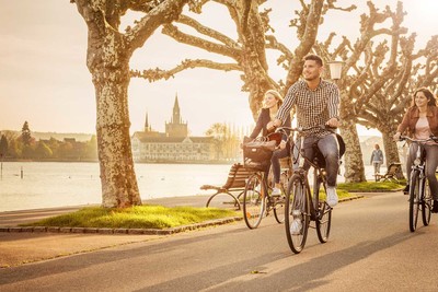 Zwei Frauen und ein Mann fahren mit ihren Fahrrädern am Ufer des Bodensees entlang. Am Ufer stehen große Bäume. Im Hintergrund sieht man die Konstanzer Altstadt.