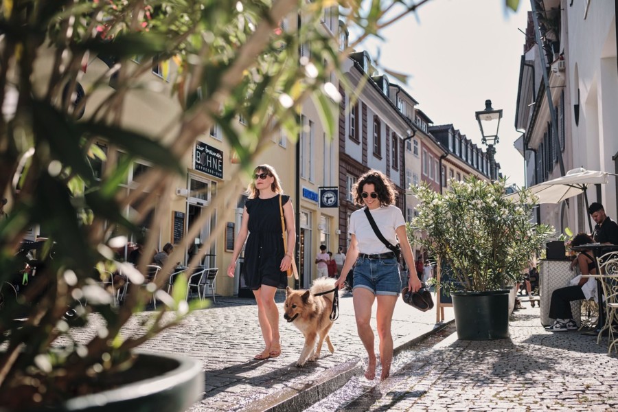 Zwei Frauen und ein Hund schlendern durch die Gassen einer Innenstadt. Ein der Frauen läuft barfuß durch einen kleinen Kanal.