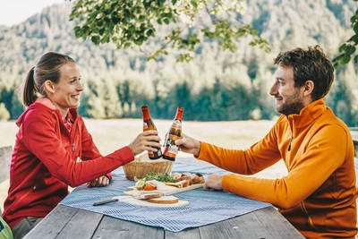 Ein Mann und eine Frau sitzen gegenüber an einem Tisch und prosten sich mit einem Bier zu. Auf dem Tisch liegt ein Vesperbrett mit Brot und Aufstrich.