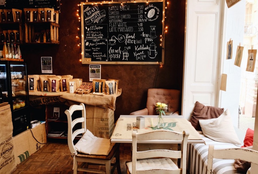 Innenraum eines Cafés. An den Wänden stehen Regale, in denen verschiedene Beutel mit Kaffee liegen. Auf der rechten Seite steht ein weißer Tisch mit Stühlen.