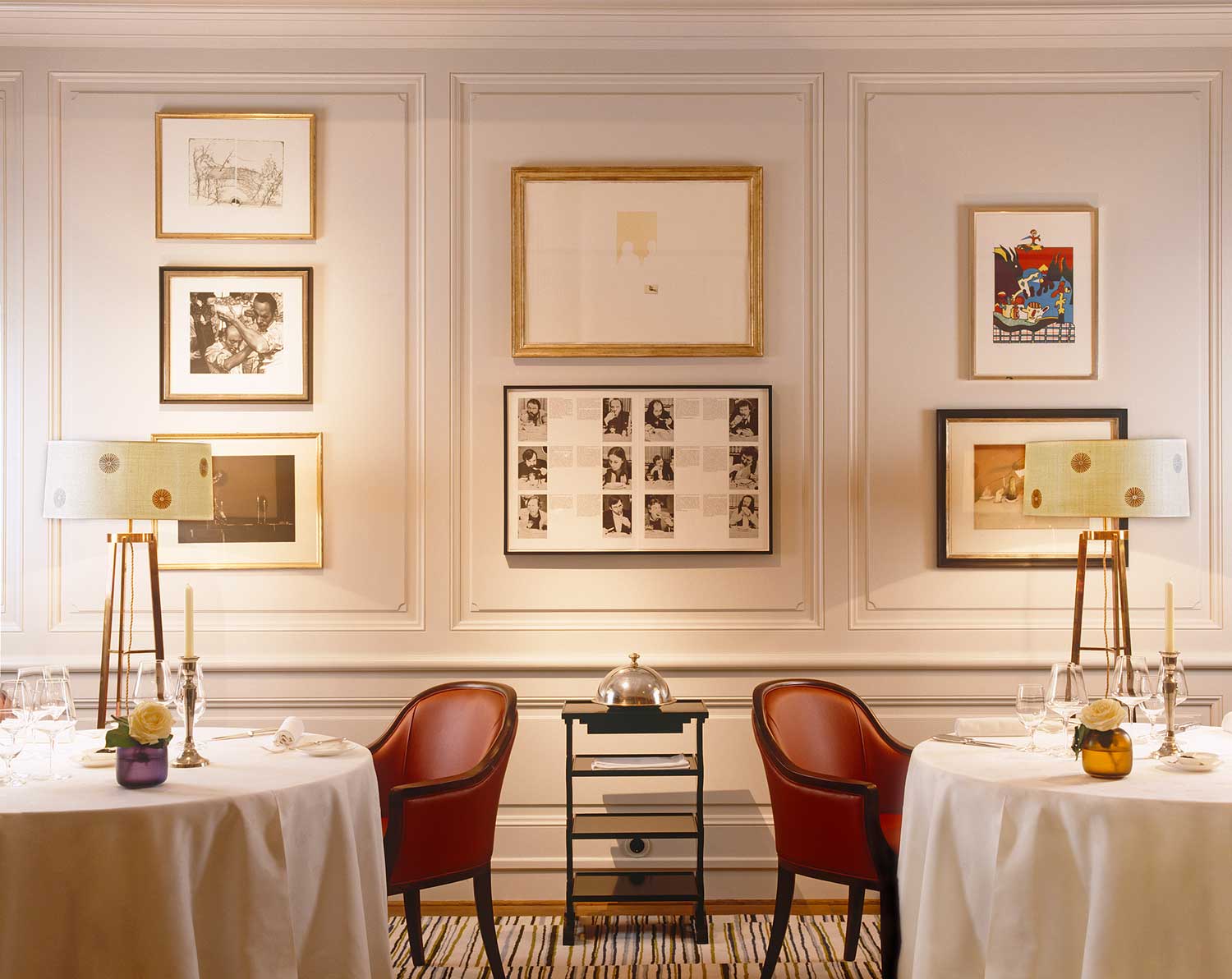 Restaurant Erbprinz. An einer weißen Wand hängen viele Gemälde. Davor stehen zwei edel gedeckte Tische mit Weingläsern und Kerzen. Neben den Tischen stehen zwei rote bequeme Stühle.