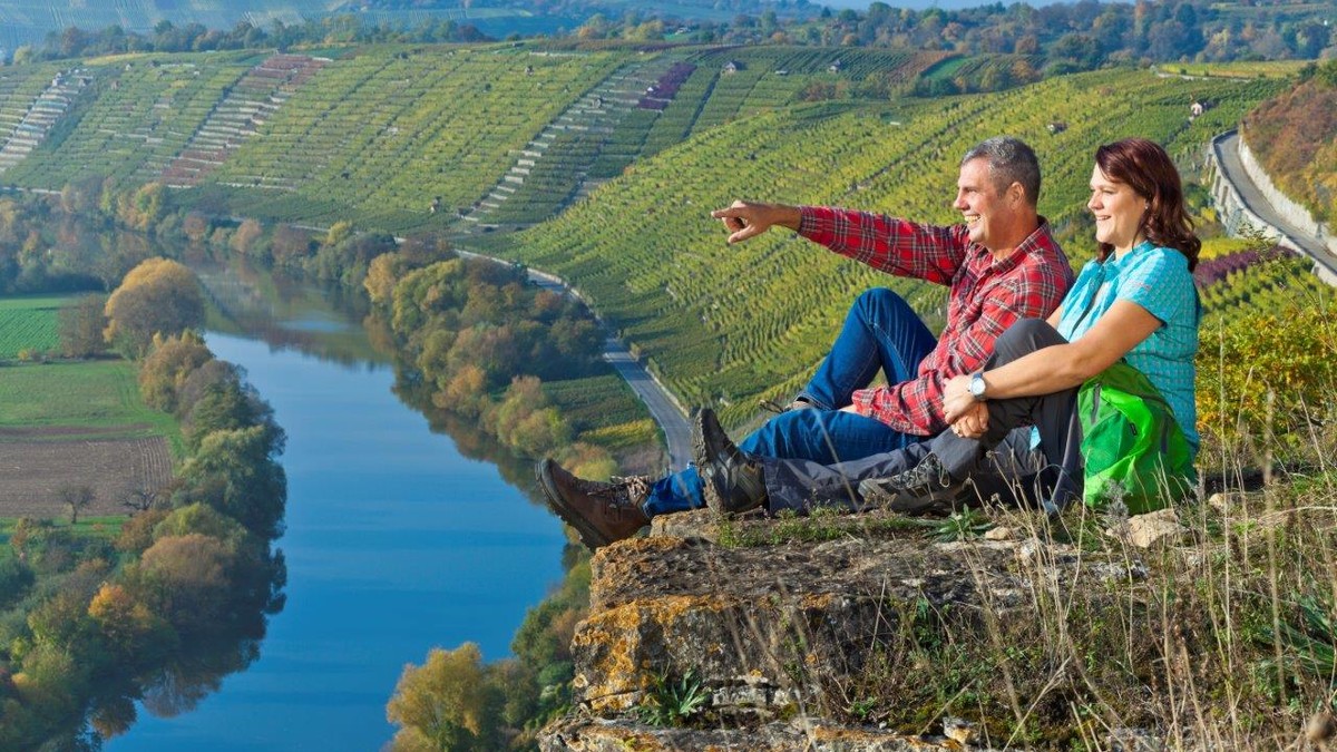 Auf der linken Seite fließt der Neckar. Rechts davon gehen Steillagen hoch, auf denen Wein angebaut wird. Auf einem Felsen am Rand des Hanges sitzen zwei Personen und schauen in die Ferne. Der Mann zeigt mit seinem Finger auf einen bestimmten Punkt in der Landschaft.