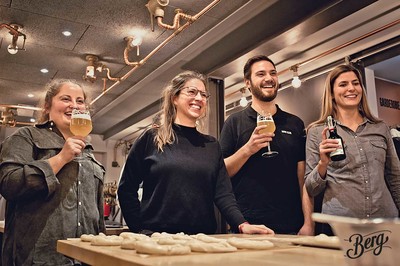 Hinter einem Holztisch stehen drei Frauen und ein Mann. Alle lachen und halten ein Bier in den Händen. Vor ihnen auf dem Tisch liegen Brezeln aus noch rohem Teig.
