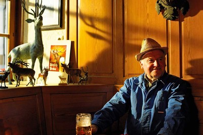 Im Inneren eines gemütlichen Restaurants sitzt ein älterer Mann auf einer Bank. Die Wände sind holzvertäfelt. Vor ihm steht ein Krug mit Bier, der Mann hat einen Strohhut auf und eine blaue Arbeitsjacke.