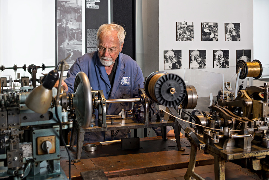 Ein älterer Mann sitzt mit einem blauen Arbeitshemd vor einer Kettenmaschine im Technischen Museum. Um ihn herum stehen noch viele andere Geräte. Im Hintergrund hängen schwarz-weiße Fotografien an der Wand.