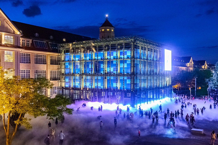 Ein viereckiges Museumsgebäude ist blau beleuchtet. Davor stehen viele Menschen.
