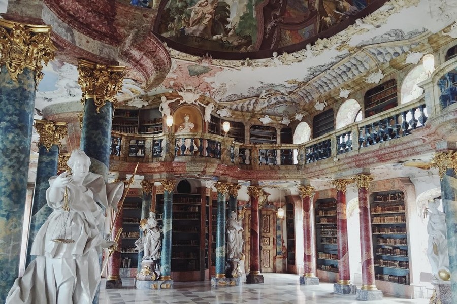 Bibliothekssaal im Kloster Wiblingen mit hohen Säulen und Bücherregalen und kunstvollen barocken Schmuckelementen.