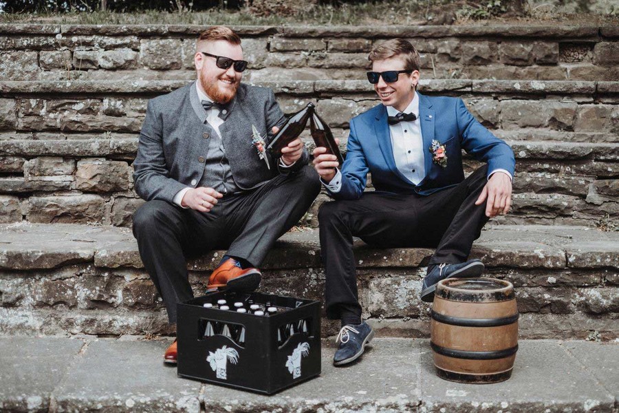 Auf einer Treppe aus Stein sitzen zwei Männer und halten eine Bierflasche in der Hand. Beide haben einen schicken Anzug an und eine Sonnenbrille auf. Der linke Mann hat sein Fuß auf einem Bierkasten abgestellt, der rechte Mann seinen Fuß auf einem kleinen Bierfass.