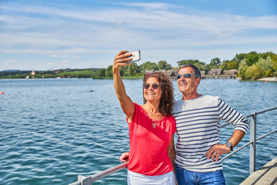 Ein Mann und eine Frau stehen eng beieinander auf einem Steg im Wasser und machen ein Selfie von sich. Im Hintergrund ist der Bodensee.