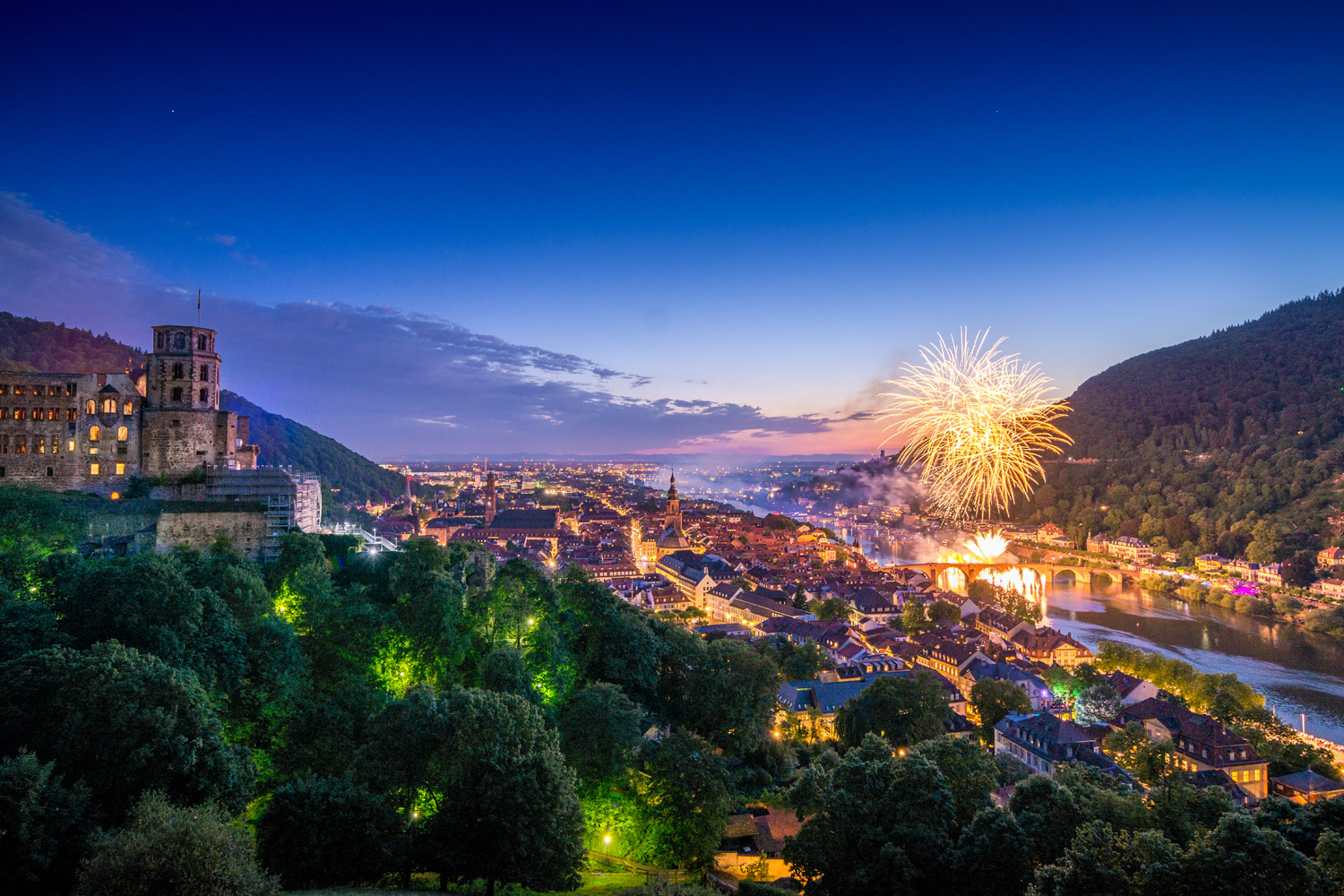 Panoramablick auf Heidelberg bei Nacht mit beleuchtetem Schloss. Auf der rechten Seite ist ein großes Feuerwerk über dem Fluss.