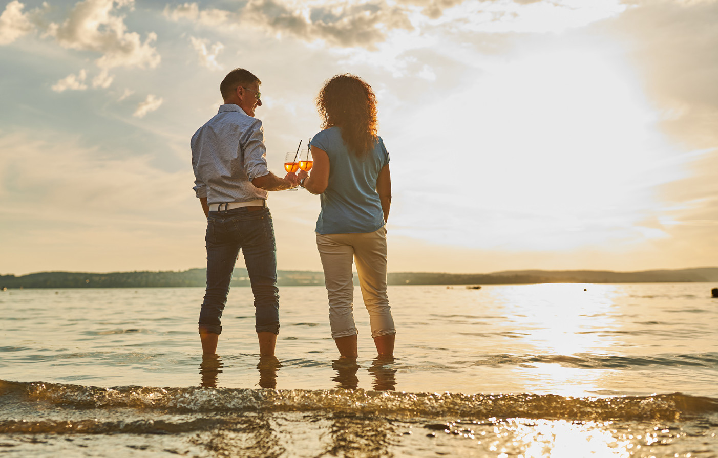Ein Mann und eine Frau stehen knöcheltief im Wasser des Bodensees und stoßen mit einem Glas gegenseitig an.