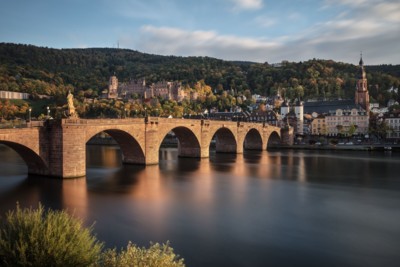 Blick auf die alte Brücke von Heidelberg bei Abenddämmerung. Im Vordergrund fließst der Neckar. Das Schloss und die Stadt Heidelberg bilden die Kulisse im Hintergrund.