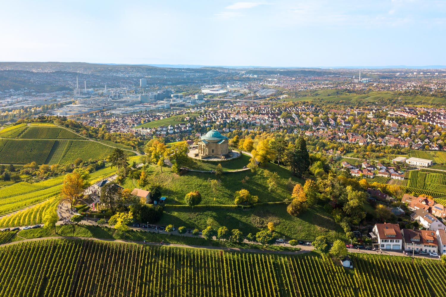 Auf einem kleinen Berg steht eine Kapelle eingerahmt von Wiesen, Feldern und Bäumen. Im Hintergrund sieht man die Hausdächer von Stuttgart.