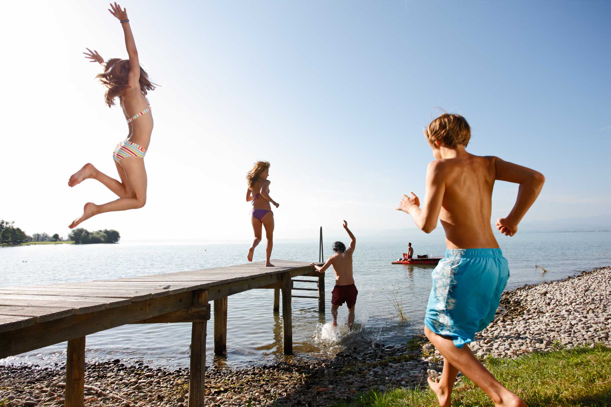 Kinder springen in Immenstaad am Bodensee ins Wasser. Zwei Kinder sind auf einem Steg aus Holz. Ein rotes Tretboot schwimmt im Wasser.