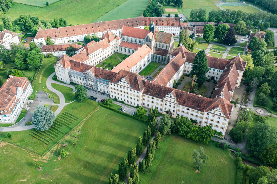 Luftaufnahme des Schloss Salem am Bodensee. Das Schloss besteht aus vielen langen Gebäuden. Um das Schloss sind Wiesen und Bäume.