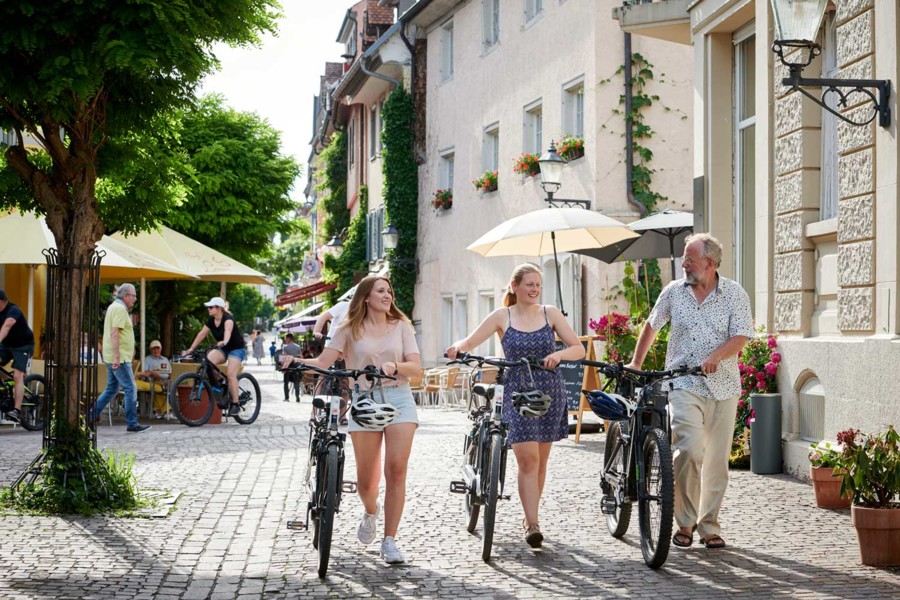 Das Bild zeigt Fahrradfahrer, die - ihr E-Bike neben sich her schiebend - durch die Altstadt von Radolfzell am Bodensee bummeln.