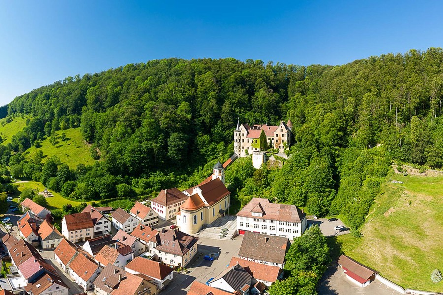 Panoramablick auf Schloss Weißenstein, wie es in einem angrenzenden grünen Mischwald liegt. Der Himmel ist strahlend blau. Zu Füßen des Schlosses liegt ein malerisches Dorf. 