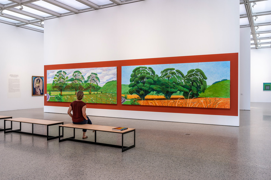 Innenaufnahme des Museums. Eine Frau sitzt lässig auf einer Sitzbank und betrachtet die Bilder an der Wand vor sich. Die zwei riesigen Bilder sind mit einem roten Hintergrund hinterlegt und es sind Bäume darauf zu erkennen, welche unterschiedlich gestaltet worden sind.