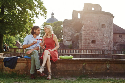 Vor dem Heidelberger Schloss sitzen ein Mann und eine Frau auf einer Mauer. Sie schauen sich verliebt an und stoßen mit zwei Weingläsern an.
