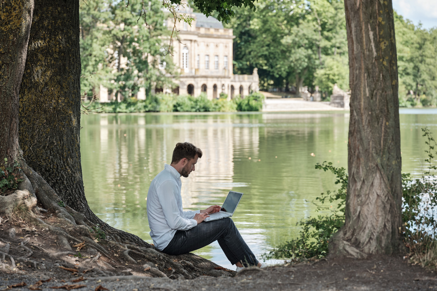 Ein Mann sitzt an einen Baum gelehnt am Ufer eines kleinen Sees und arbeitet an einem Laptop. Im Hintergrund befindet sich ein Schloss.