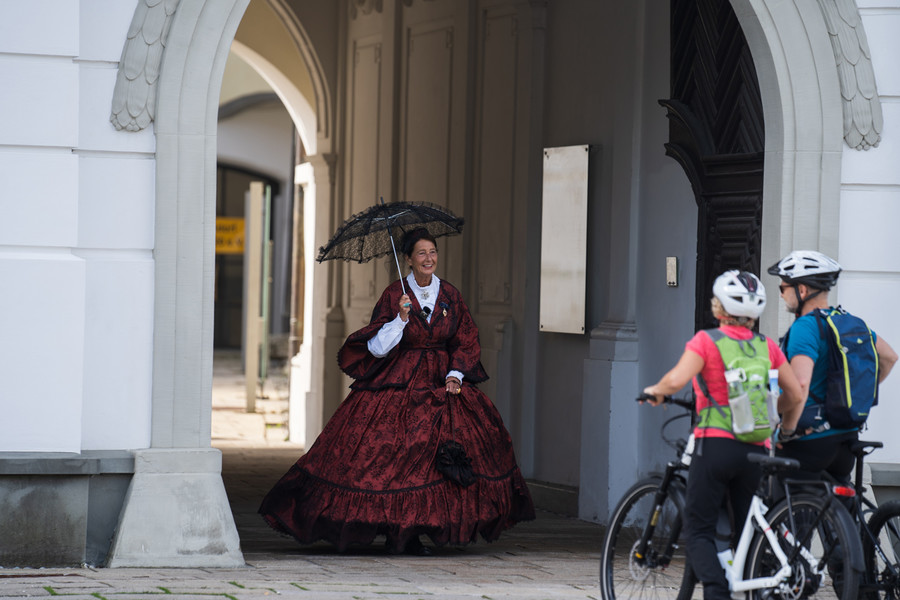 Die zwei fahrradfahrenden Personen werden im Schloss Aulendorf von einer verkleideten Frau in einem roten Kleid mit einem schwarzen Schirm in der Hand empfangen. 