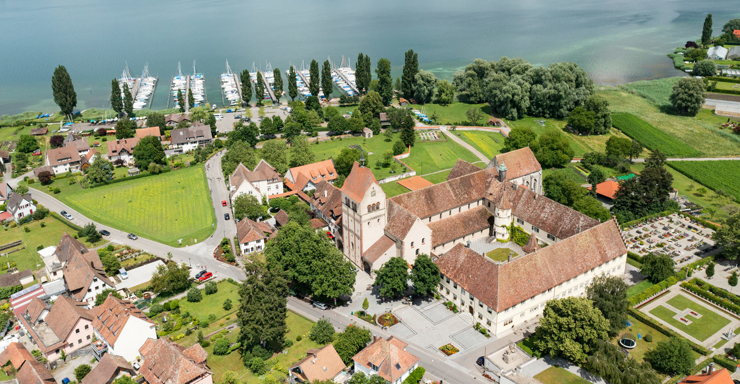 Luftaufnahme der Klosteranlage Reichenau. Die Anlage ist von Wohnhäusern umgeben und im Hintergrund ist der Bodensee.