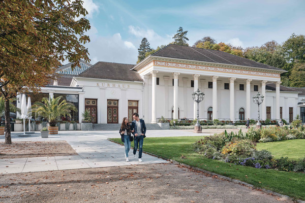 Vor dem Kurhaus in Baden-Baden laufen ein Mann und eine Frau. Das Kurhaus ist weiß und hat vor dem Eingang viele hohe Säulen.