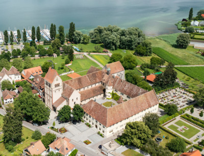 Luftaufnahme der Klosteranlage Reichenau. Die Anlage ist von Wohnhäusern umgeben und im Hintergrund ist der Bodensee.