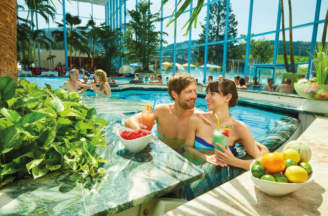 Ein Mann und eine Frau lehnen sich in einem Schwimmbecken an den Rand und trinken Cocktails. Im Hintergrund schwimmen andere Menschen.