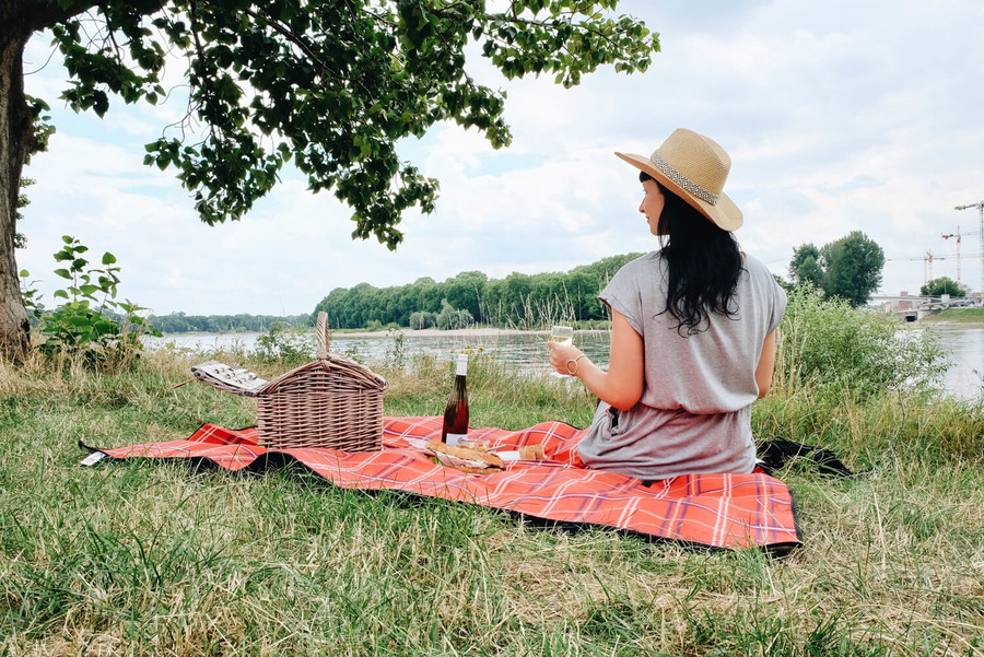 Auf einer roten Picknickdecke sitzt eine Frau auf einer Wiese am Rhein und trinkt einen Wein.