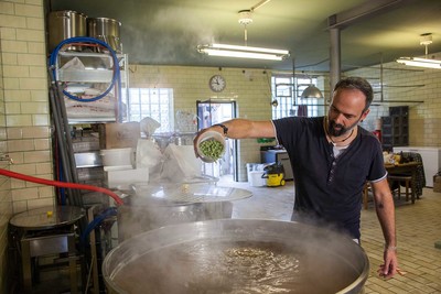 Ein Mann steht in einer Brauerei vor einem großen Kessel in dem Bier gebraut wird und aus dem es herausdampft. 