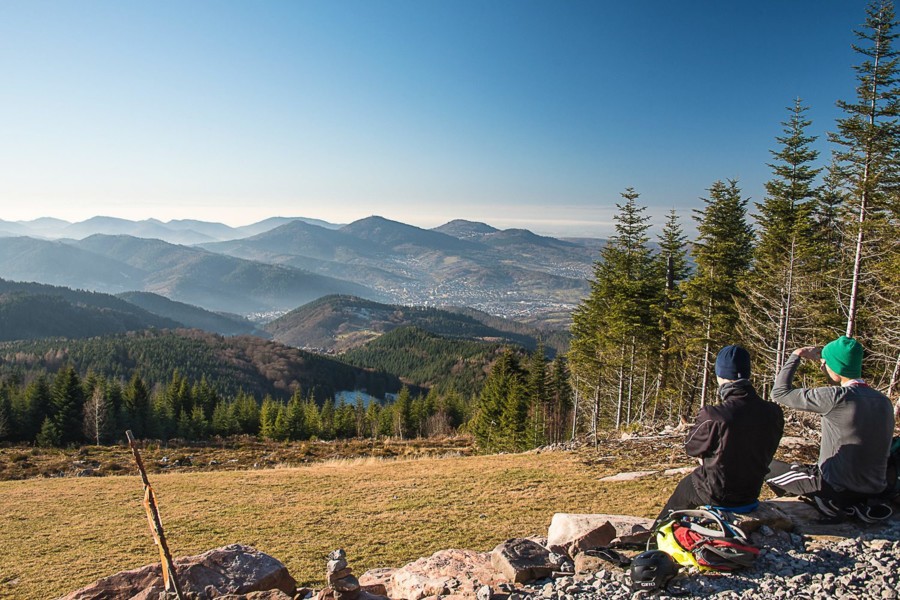 Zwei Wanderer sitzen an einem Aussichtspunkt und genießen die Aussicht in ein bewaldetes Tal.