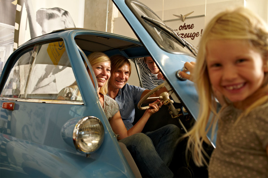 Zwei Erwachsene sitzen in einem kleinen blauen Oldtimer. Ein Mädchen öffnet ihnen lachend die Autotür.